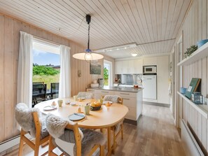 Une pièce chaleureuse et bien éclairée, avec une table à manger en bois dressée pour le repas, une cuisine équipée d’appareils modernes et une vue sur la verdure environnante.