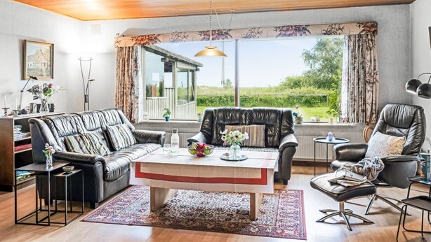 Amplia sala de estar con techo de madera, un ventanal con vistas a la vegetación, un sofá de cuero negro, una mesa de centro y un sillón negro.