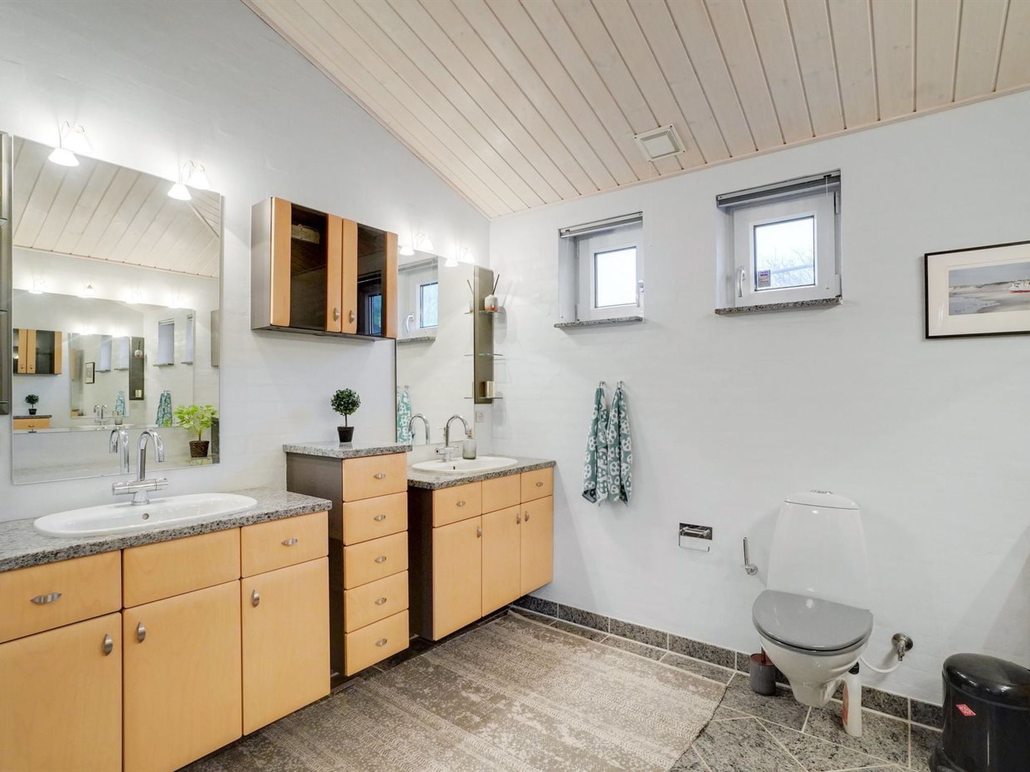 Un baño moderno con dos lavamanos, un inodoro y una zona de ducha. Hay ventanas que proporcionan luz natural y un espejo grande sobre los lavamanos.