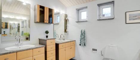 Un baño moderno con dos lavamanos, un inodoro y una zona de ducha. Hay ventanas que proporcionan luz natural y un espejo grande sobre los lavamanos.