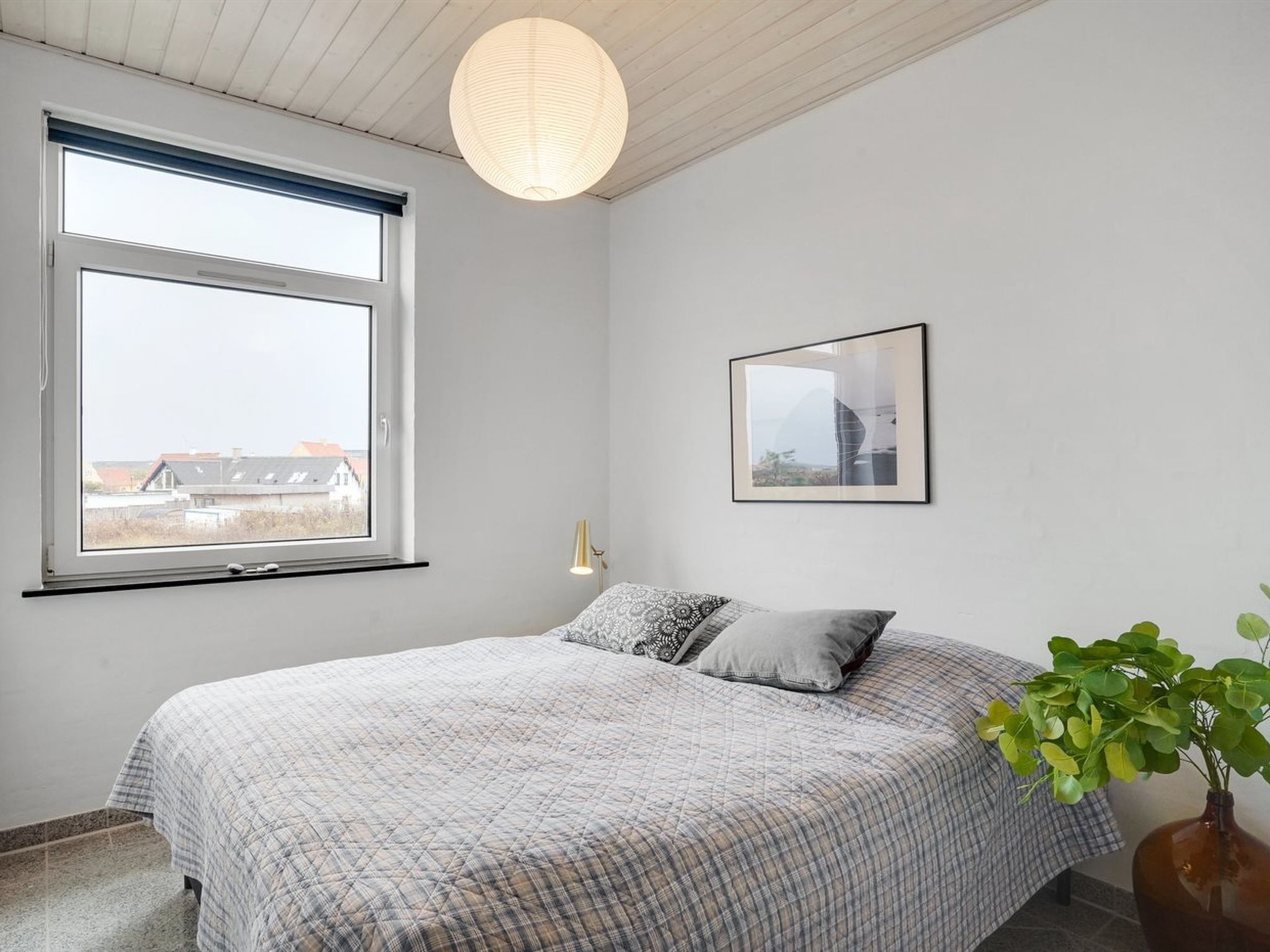 Un dormitorio con un ventanal grande, una cama con colcha a cuadros, un cuadro en la pared y una planta en maceta.