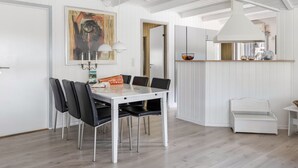 A modern dining area with a white table and chairs, a painting on the wall, and a kitchen area in the background.