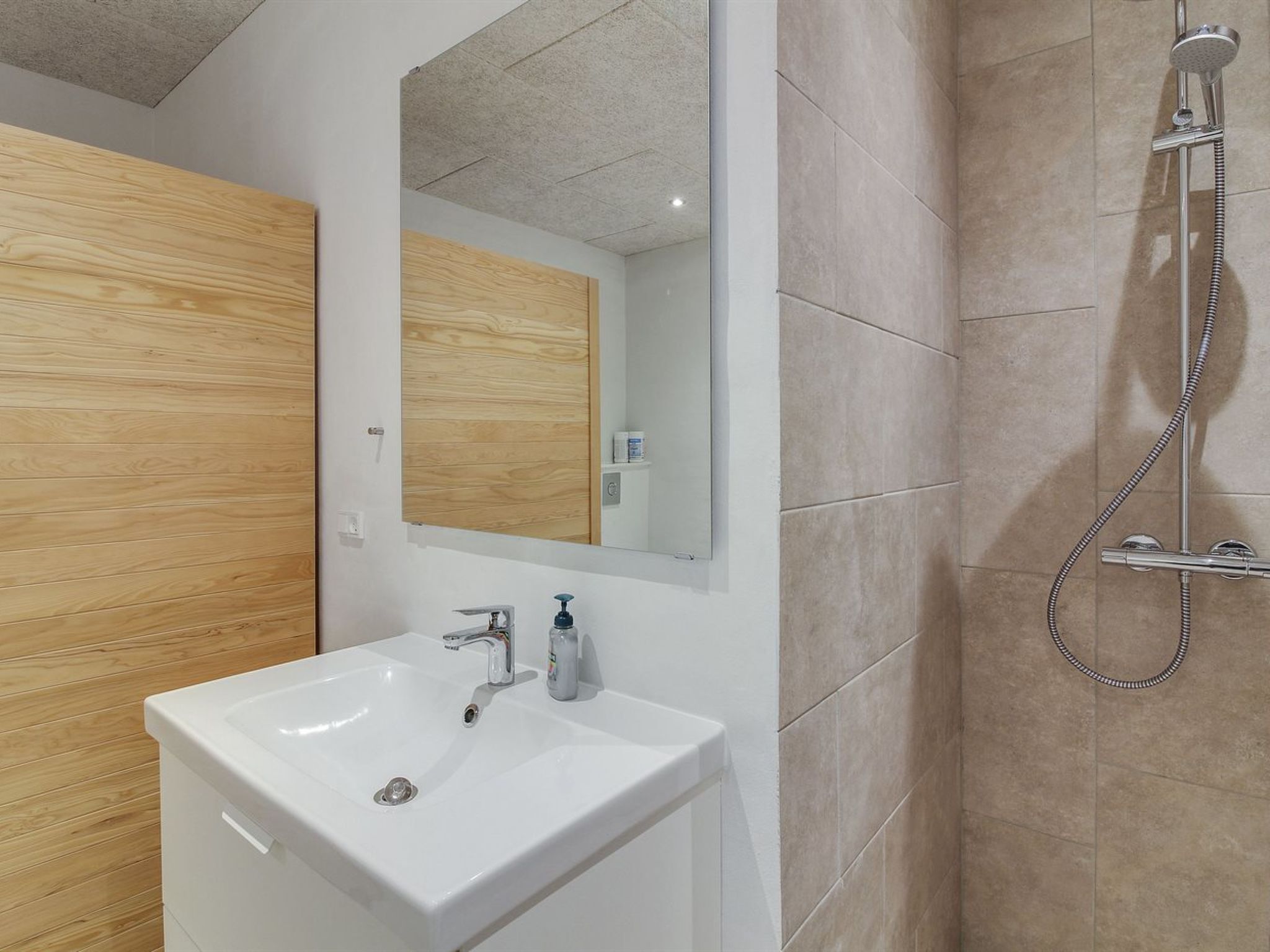 A modern bathroom with a white sink, a wooden door, a large mirror, and a shower area.