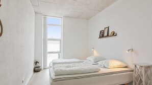 Ein schlichter Schlafzimmer mit großem Fenster, einem Bett mit weißer Bettwäsche, einem kleinen Nachttisch und einer Wandablage mit Dekoration.