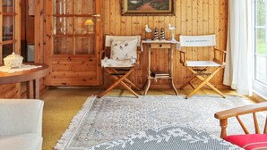Una habitación acogedora con paredes de madera, un cuadro enmarcado, una mesita con una lámpara y dos sillas plegables.
