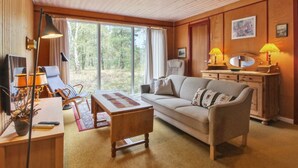 Una sala de estar acogedora con un sofá, una mesita de centro y un escritorio con una lámpara. Amplias ventanas ofrecen vistas al exterior.