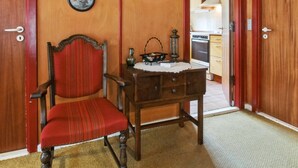 Una habitación con puerta de madera, un sillón tapizado en rojo, una mesa de madera con mantel de encaje, y una cocina visible a través de un vano abierto.