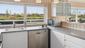 Une cuisine moderne équipée d’un lave-vaisselle, d’un four encastré et d’un grille-pain. On y trouve trois lampes suspendues, une fenêtre donnant sur un paysage verdoyant et des maisons, ainsi qu’un plan de travail agrémenté d’une plante en pot.