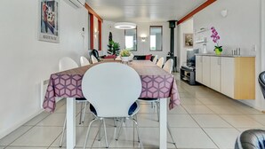 Un comedor moderno con una mesa larga, sillas blancas y un mantel estampado. Se observa una cocina con armarios y una estufa a la derecha.