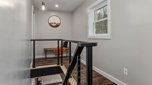 Una escalera moderna con barandilla de metal negro y peldaños de madera, que conduce a una habitación con pared blanca y ventana.