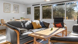 Ein modernes Wohnzimmer mit grauem Sofa, Holztisch und einem Essbereich mit Tisch und Stühlen. Große Fenster sorgen für natürliches Licht und Blick ins Grüne.