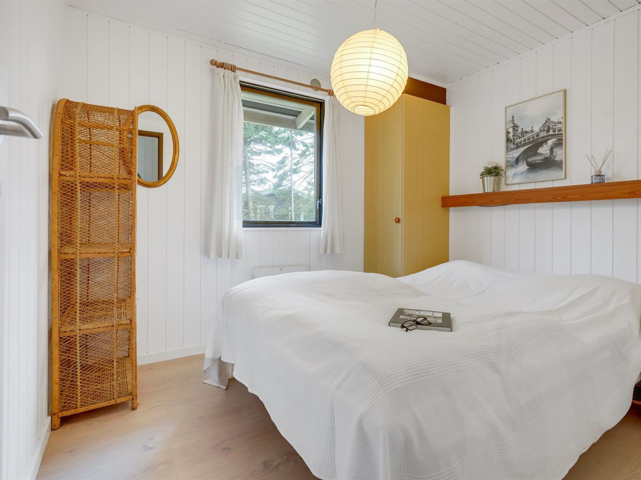 Een slaapkamer met een groot bed, een houten garderobe, een spiegel, een raam met gordijnen, een hanglamp en een ingelijst schilderij aan de muur.