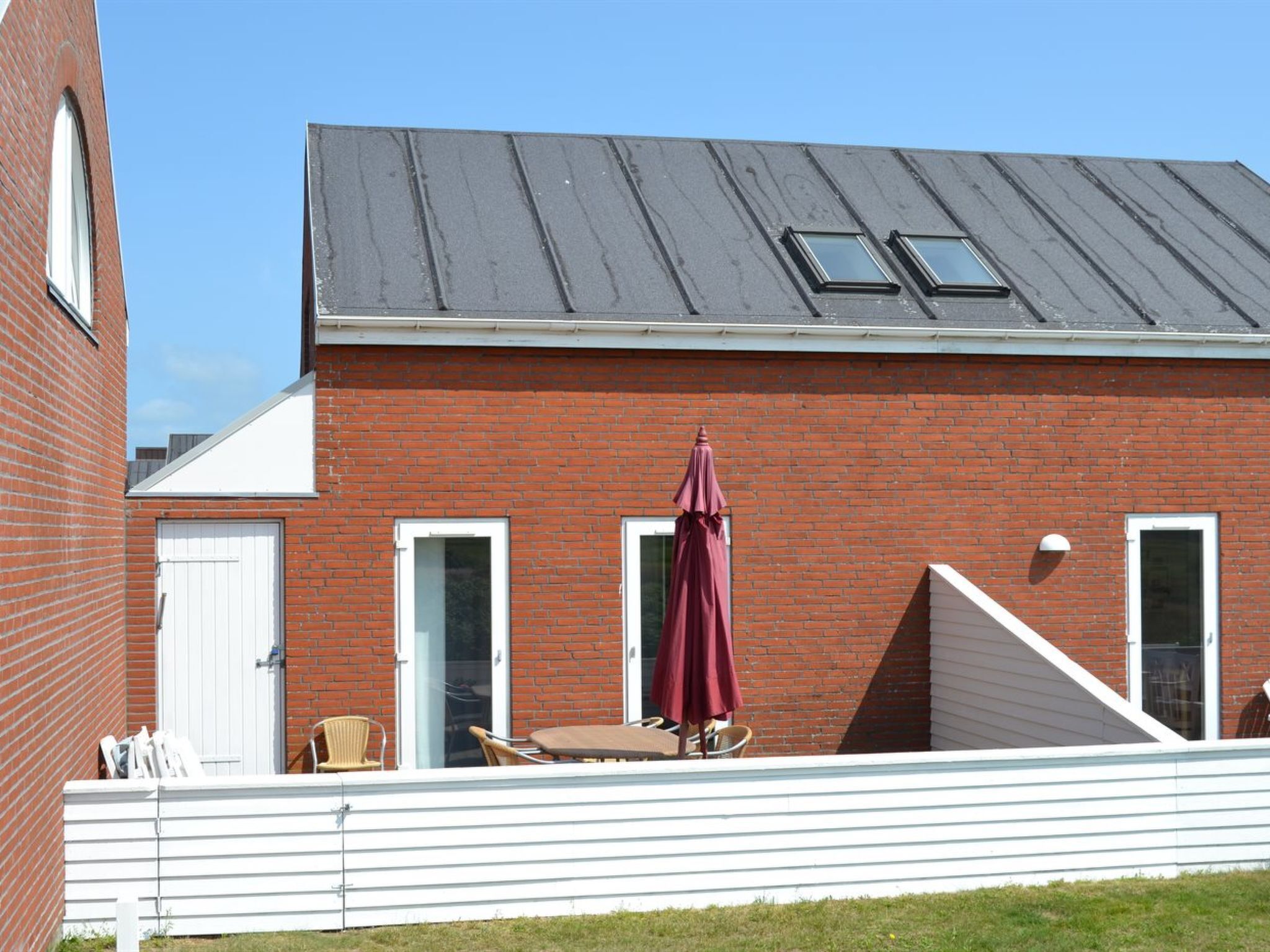 Een gebouw van rode bakstenen met een witte deur, een overdekte patio met een tafel en stoelen, en een pergola met een rode parasol.