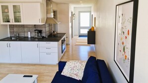 Et moderne køkken med hvide skabe, en sort bordplade og et indbygget komfur. I opholdsstuen er der en blå sofa med et blomstret pled og et indrammet botanisk tryk på væggen.