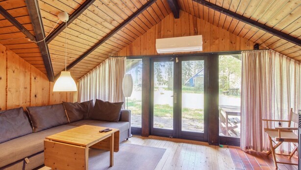 A cozy cabin interior with a wooden ceiling, a sofa, a wooden table, and a view through large glass doors.