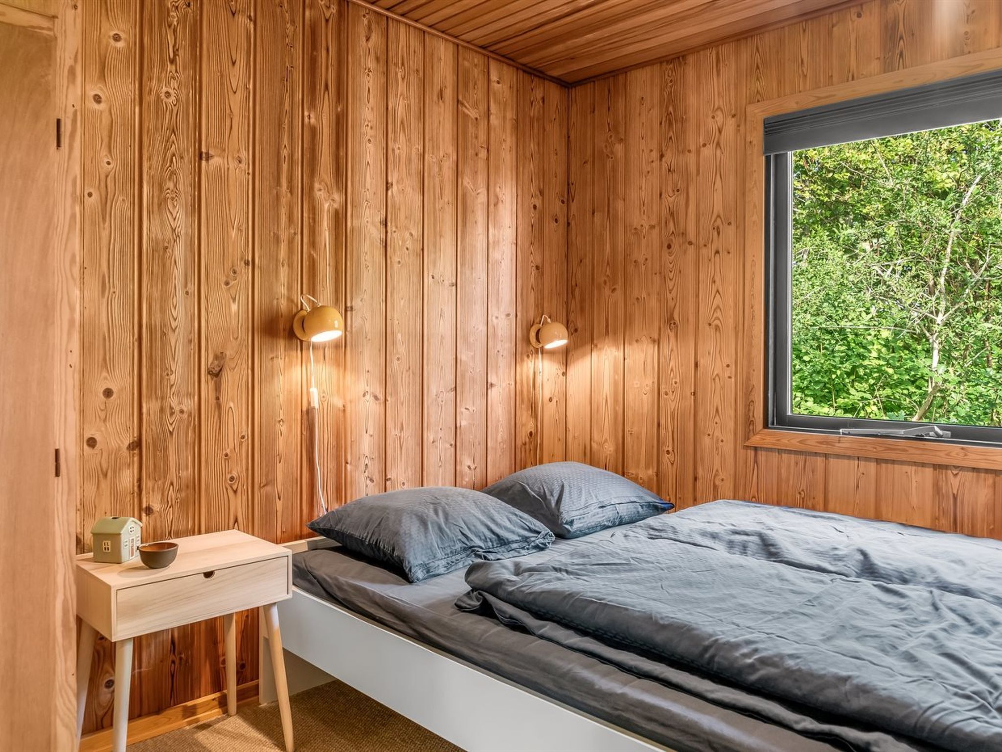 A bedroom with wooden walls, a bed with grey bedding, a bedside table with a lamp, and a window with a view of greenery.