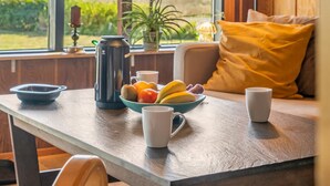 A breakfast table with a fruit bowl, coffee pot, and cups, a window with a view of greenery, and a cozy seating area with a yellow pillow. - "Tayana" - 75m to the fjord by Interhome (Storvorde)