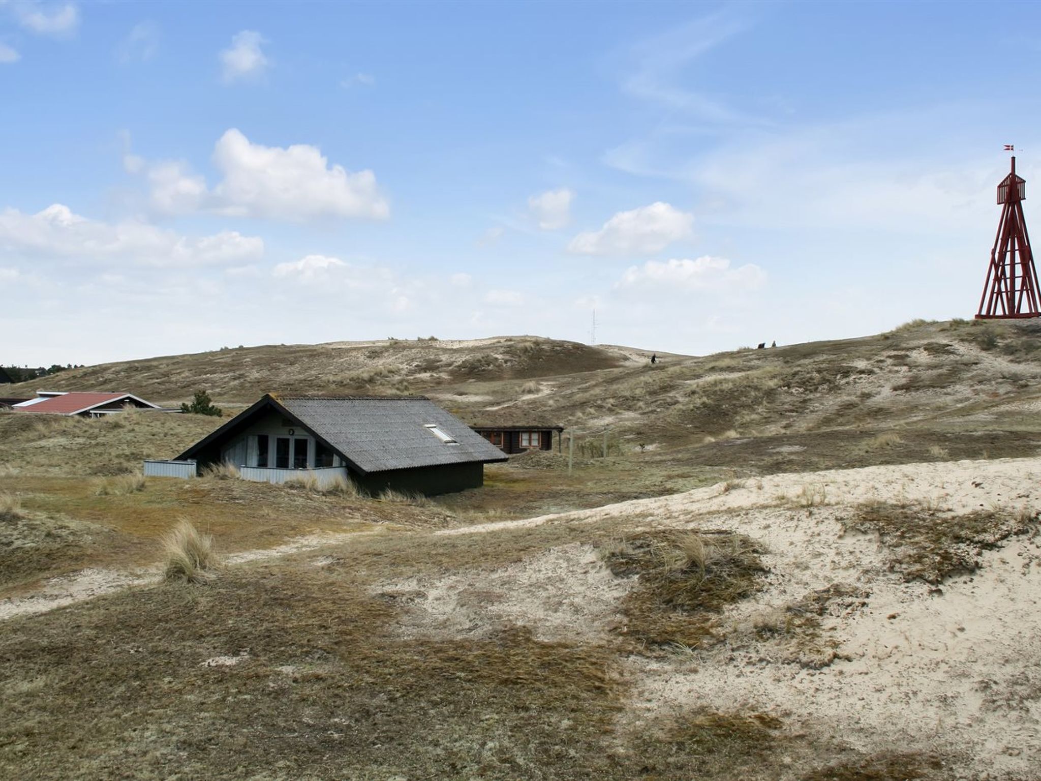 En avlägsen, enkel stuga med en grusväg som leder fram till den, omgiven av öppna fält och ett rött torn i fjärran.