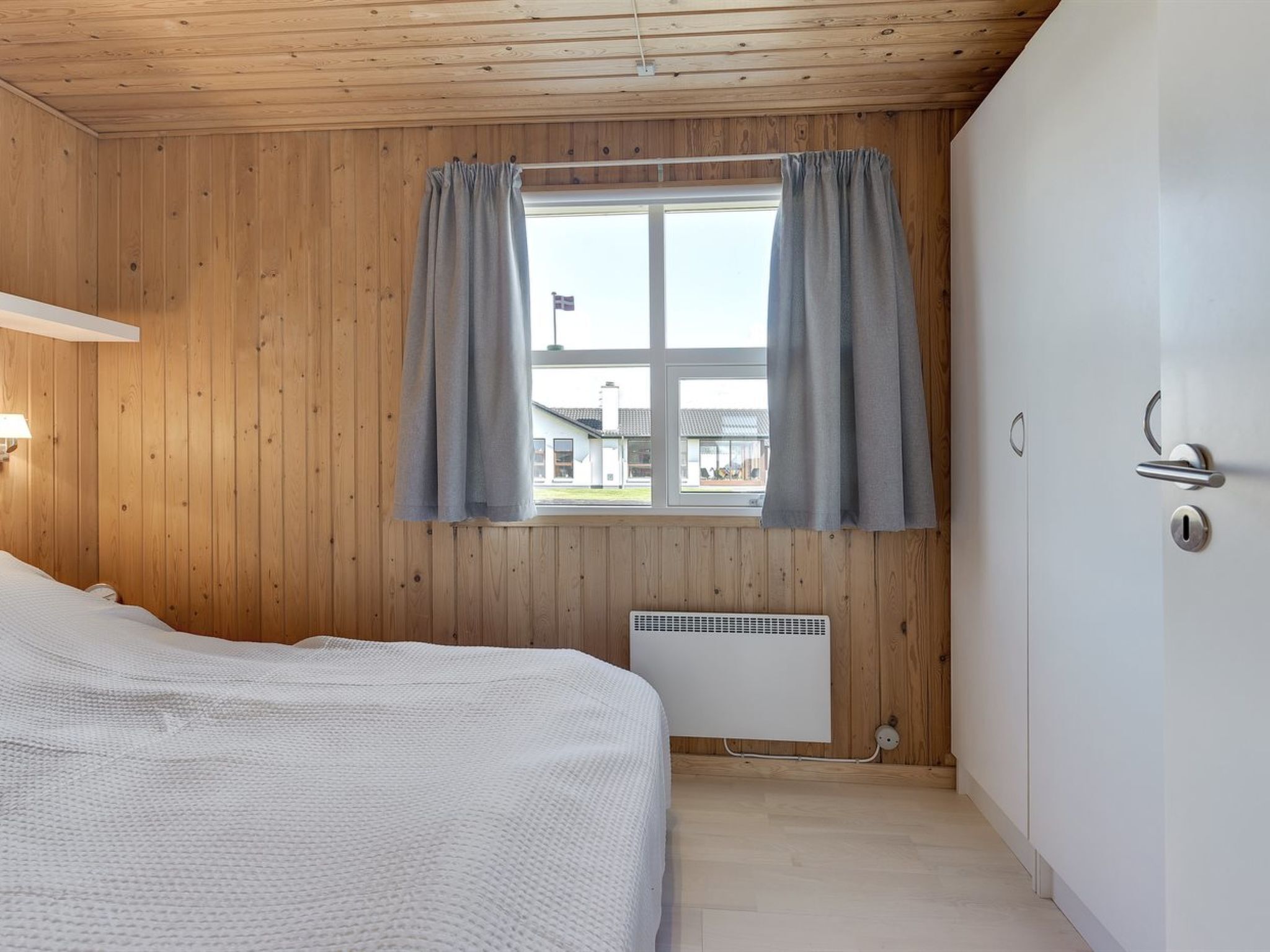 A room with wooden walls, a bed with a white cover, a window with grey curtains, and a white door with a handle.