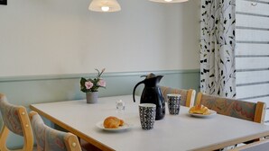 Una mesa de comedor con mantel blanco, dos lámparas colgantes, un florero, una cafetera, dos tazas estampadas, un plato con un croissant y una silla con un cojín de colores.