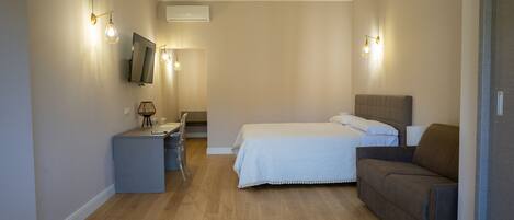 A bedroom with a bed, a desk, a chair, a sofa, and wall-mounted lighting.