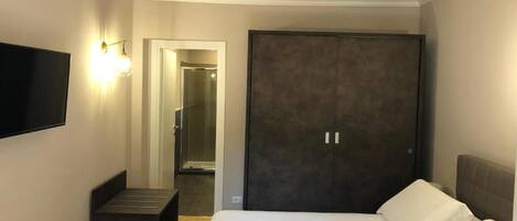 Habitación de hotel con una cama, un armario oscuro, un banco y un televisor de pantalla plana montado en la pared.