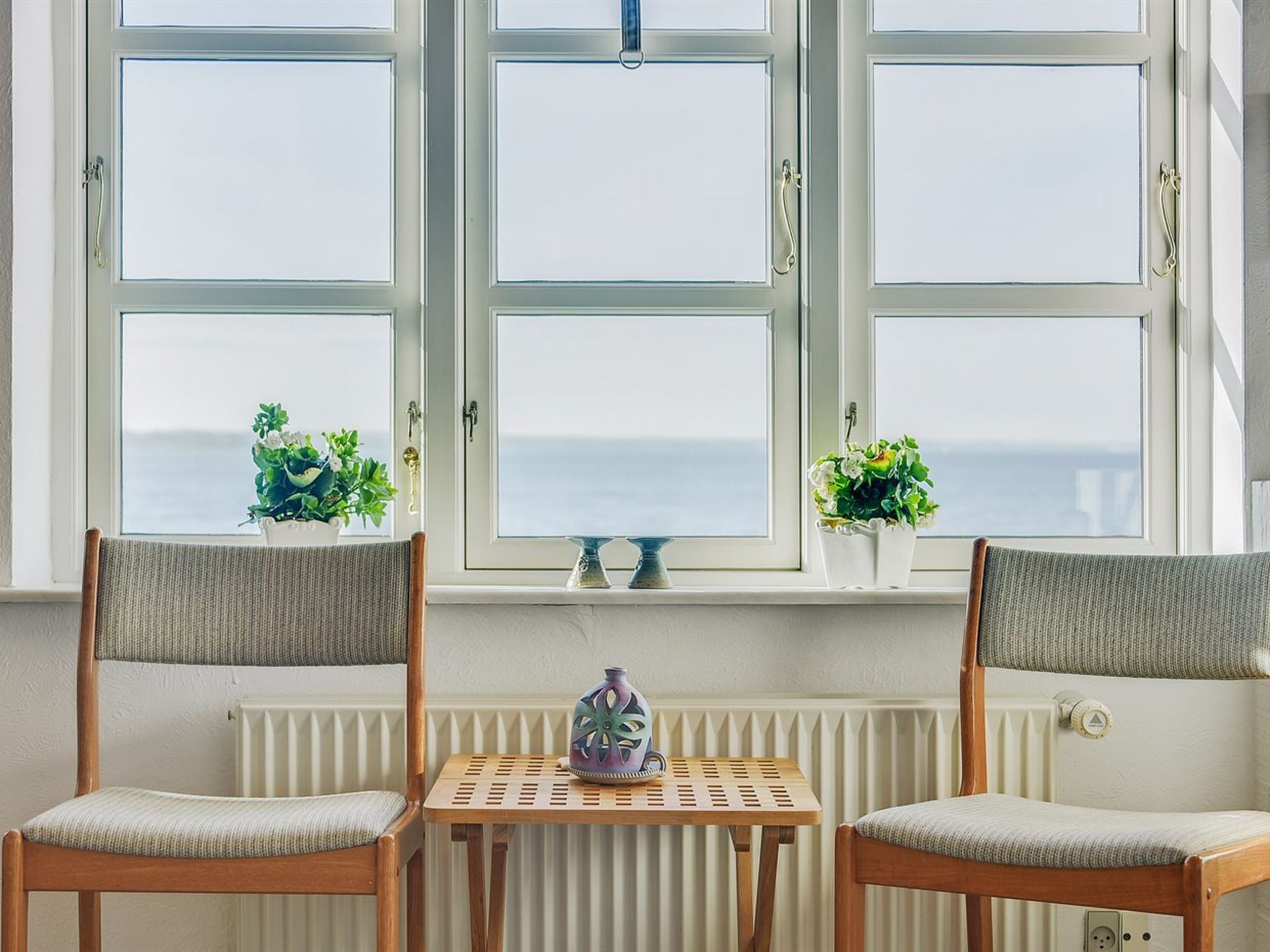 A room with two wooden chairs and a small table, a white radiator, and a window with a view of the sea.
