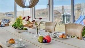 Une table à manger, dressée dans une pièce dotée de grandes fenêtres donnant sur un espace extérieur pittoresque, est garnie d’une variété de fruits, de pâtisseries et d’une théière.