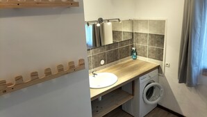 A compact bathroom with a wooden vanity, a sink, a mirror, a towel rack, a washing machine, and a shower curtain. - Maison Classée 1 Étoile (Vallouise-Pelvoux)