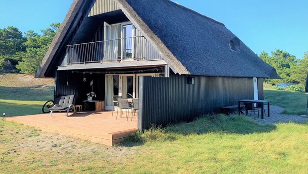 A thatched-roof house with a balcony, a covered outdoor seating area, and a grassy yard.