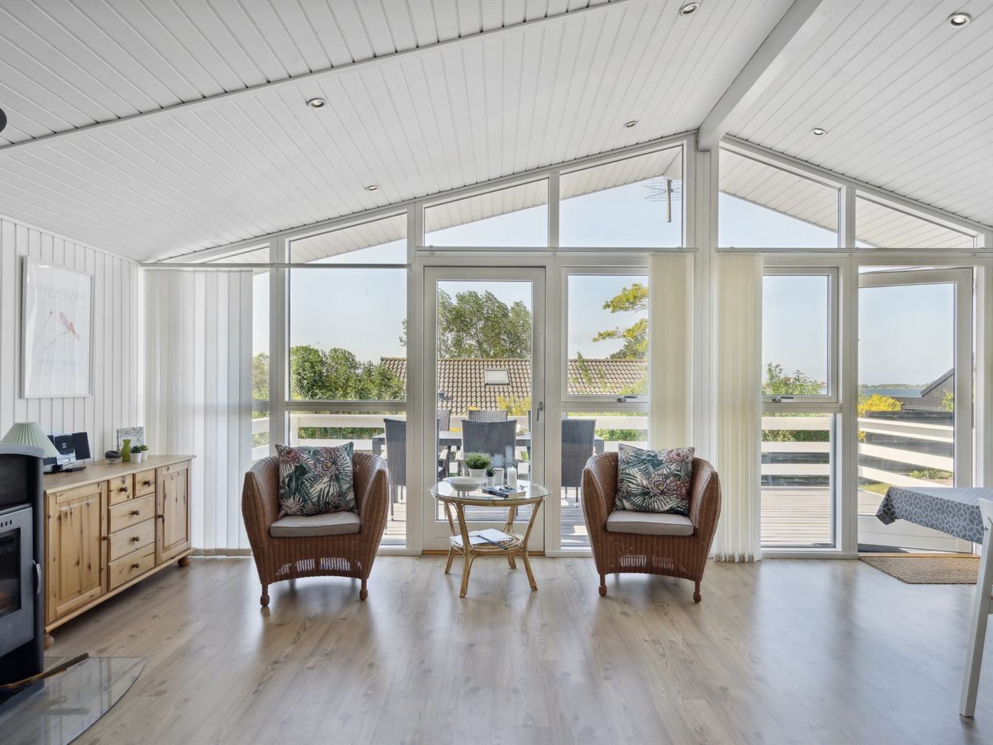 A spacious room with a high ceiling, large windows, and a view of the outdoors. There are two wicker chairs with cushions, a small table, and a cabinet with a television.
