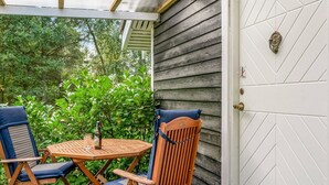 Une terrasse en bois avec une table et deux chaises, une porte blanche ornée d’un motif géométrique et un espace abrité doté d’un plafond en bois.