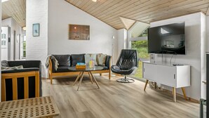 Un moderno salón con techo de madera, un sillón de cuero negro, un sofá, una mesa de centro de vidrio y un televisor de pantalla plana montado en la pared.