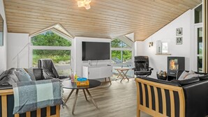 Una sala de estar moderna con televisor, sofá y chimenea, ventanas amplias y techo de madera.