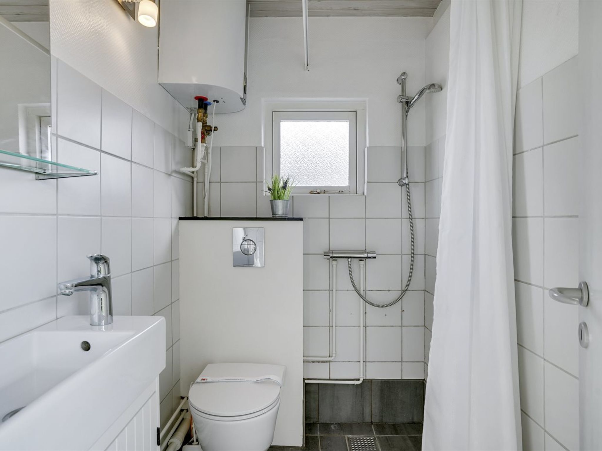 Un bany compacte amb un vàter blanc, un lavabo, una dutxa i una petita finestra.