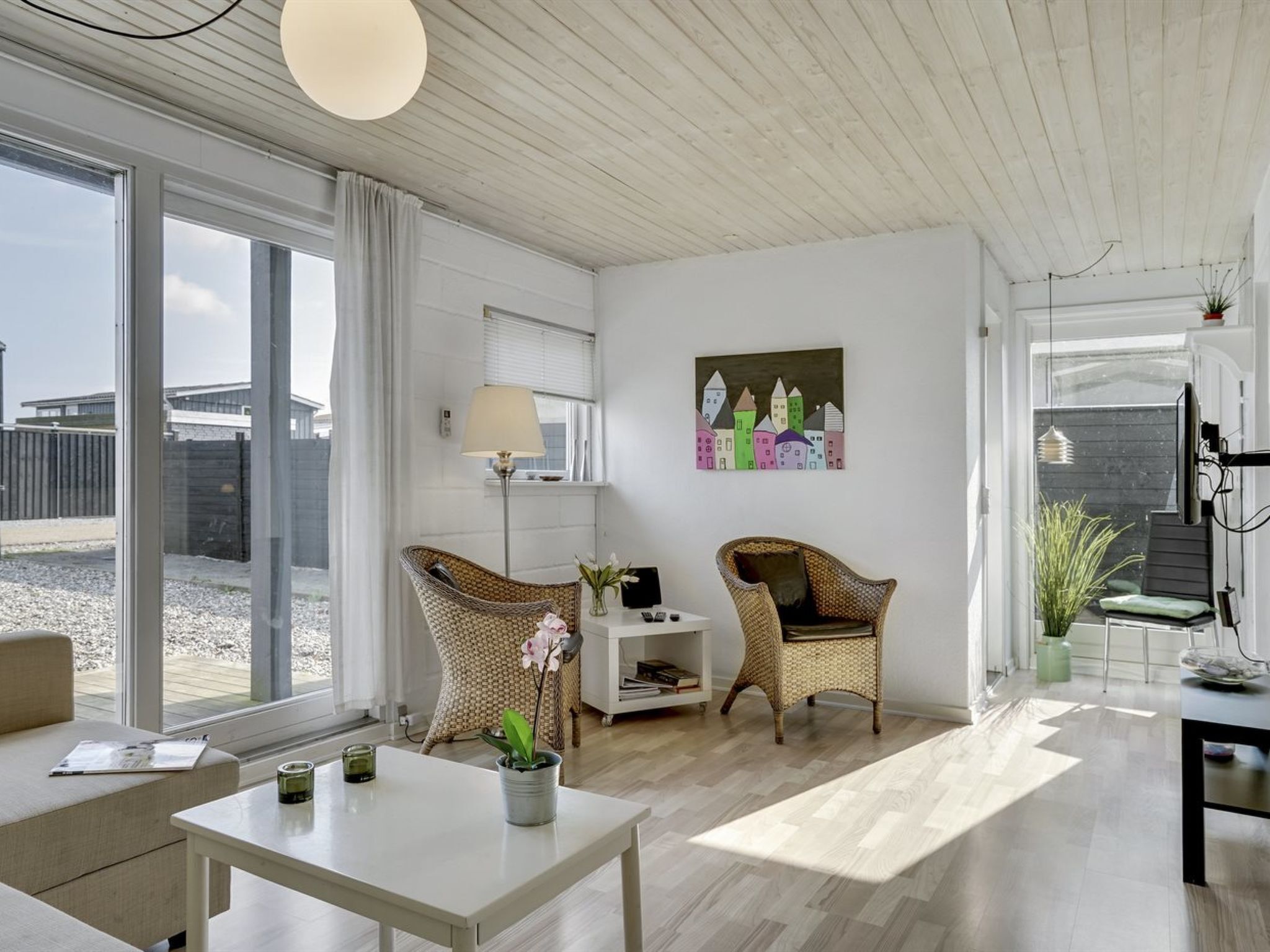 Een moderne woonkamer met een groot raam, twee rieten fauteuils, een witte tafel en een schilderij aan de muur.