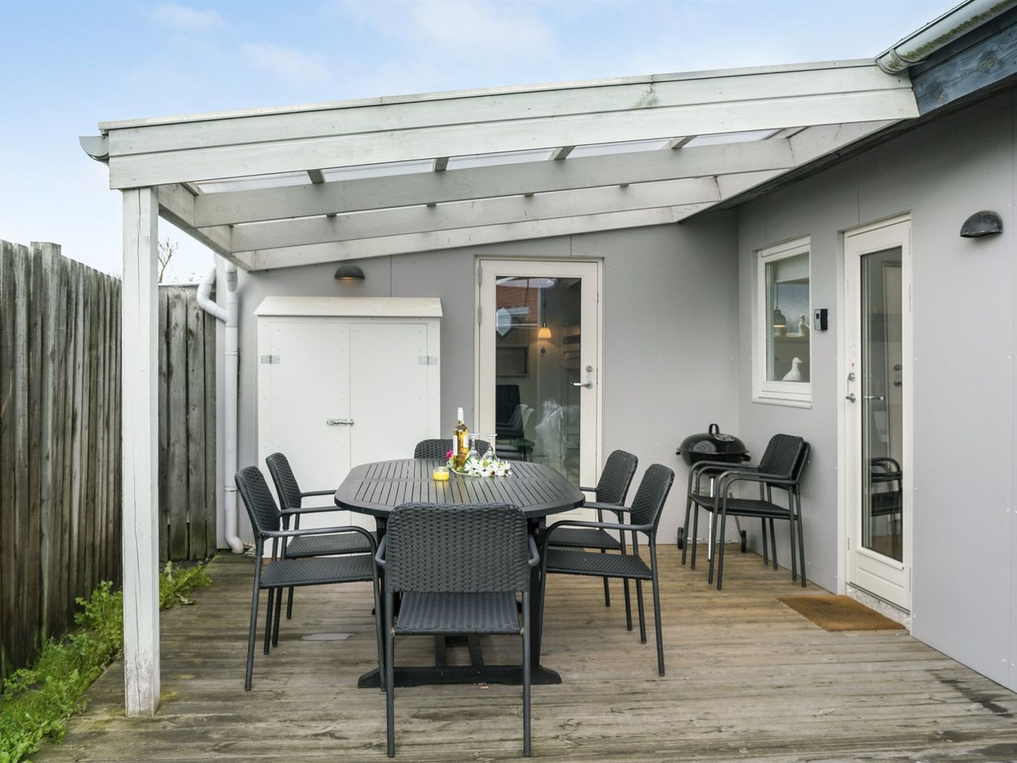Een overdekt terras met een ronde tafel en stoelen, een houten terras en een witte deur.