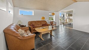 Ampla sala de estar com pé direito alto, sofás de couro, mesa de centro de madeira e vista para a cozinha.