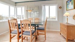 Un coin repas avec des chaises et une table en bois, de grandes fenêtres, un tableau et une lampe.