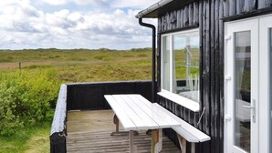 Une terrasse en bois avec une table et des chaises blanches, un mur extérieur noir et une grande fenêtre.
