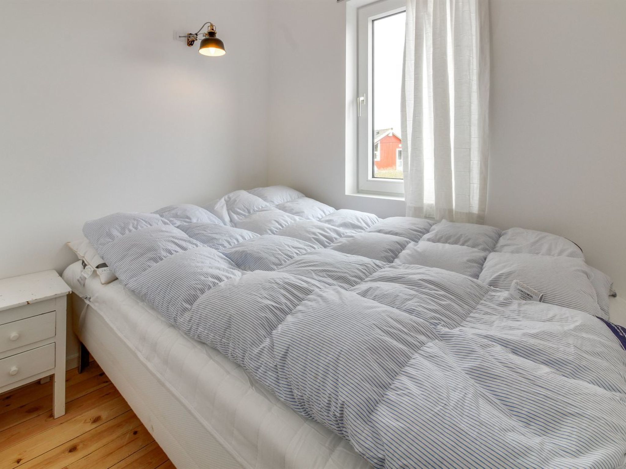 Un dormitorio con cama, mesita de noche, ventana con cortinas y lámpara de pared.