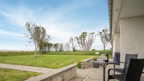 Eine Terrasse mit Tisch und Stühlen, einem Betonpflanzgefäß und Blick auf ein Wiesfeld mit Bäumen.