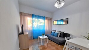 Uma sala de estar pequena com um sofá, uma TV em um suporte, uma mesinha e um lustre.