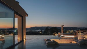 Eine moderne Außenterrasse mit Glaswand, Liegestühlen und Blick auf eine Stadt im Abendlicht.
