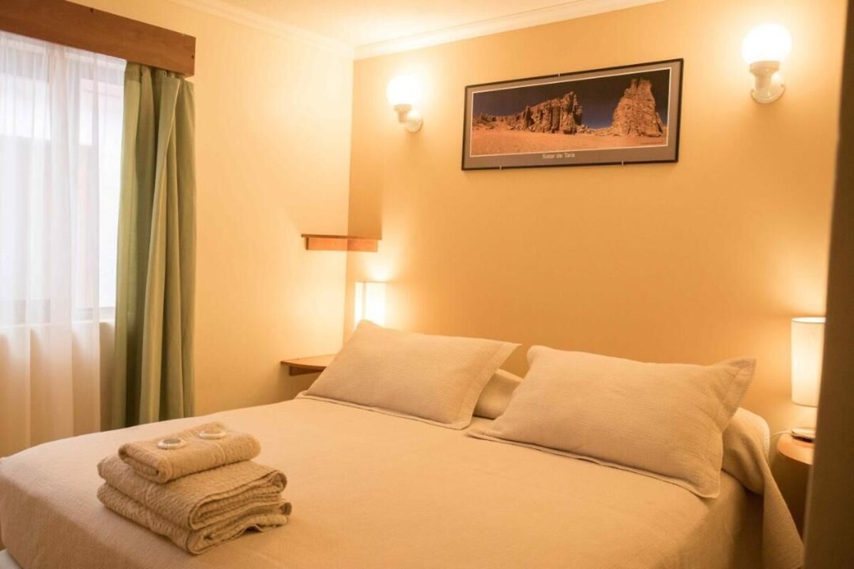 A neatly made bed with folded towels, a framed picture on the wall, and a window with sheer curtains.