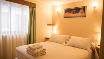 Una cama bien hecha con toallas dobladas, un cuadro en la pared y una ventana con cortinas traslúcidas.