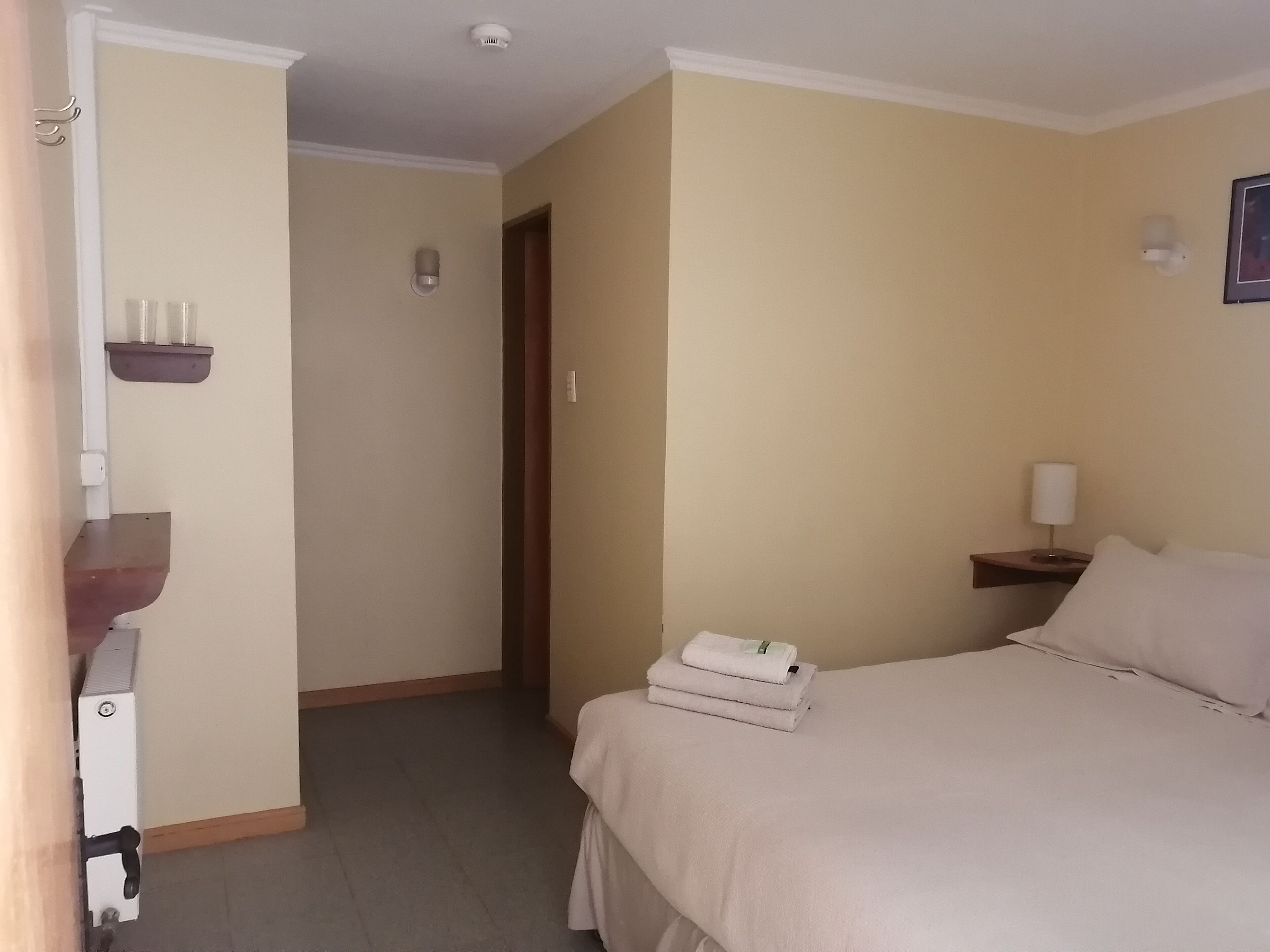 Hotellihuone, jossa on sänky, yöpöytä lampun kanssa, seinälle kiinnitetty hylly, jossa on kuppi, ja ovi, joka johtaa toiseen huoneeseen.