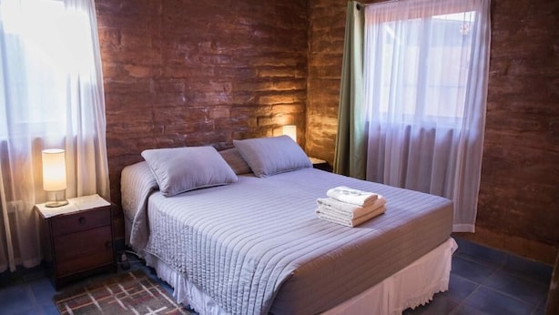 Un dormitorio con una cama grande, dos almohadas, una mesita de noche con lámpara y una ventana con cortinas traslúcidas.
