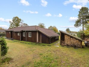 Une cabane en bois à deux étages, avec un toit en tuiles, une véranda couverte et une pile de bois de chauffage.