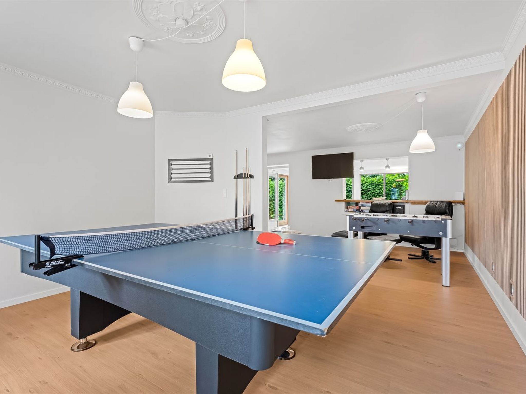 A ping pong table in a room with a wooden floor, a sofa, a TV, and pendant lights.